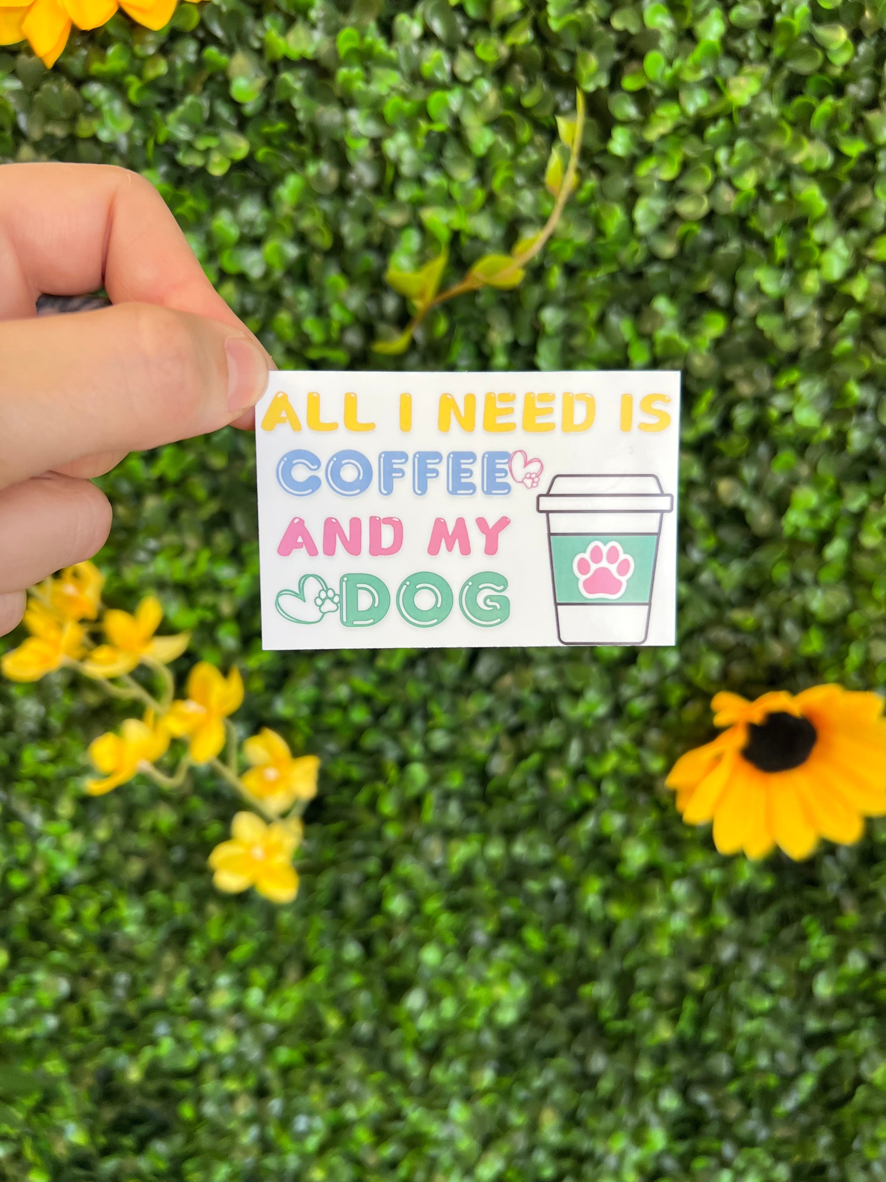 Hand holding All I Need is Coffee and my Dog Sticker with coffee cup and paw design on green leaf background