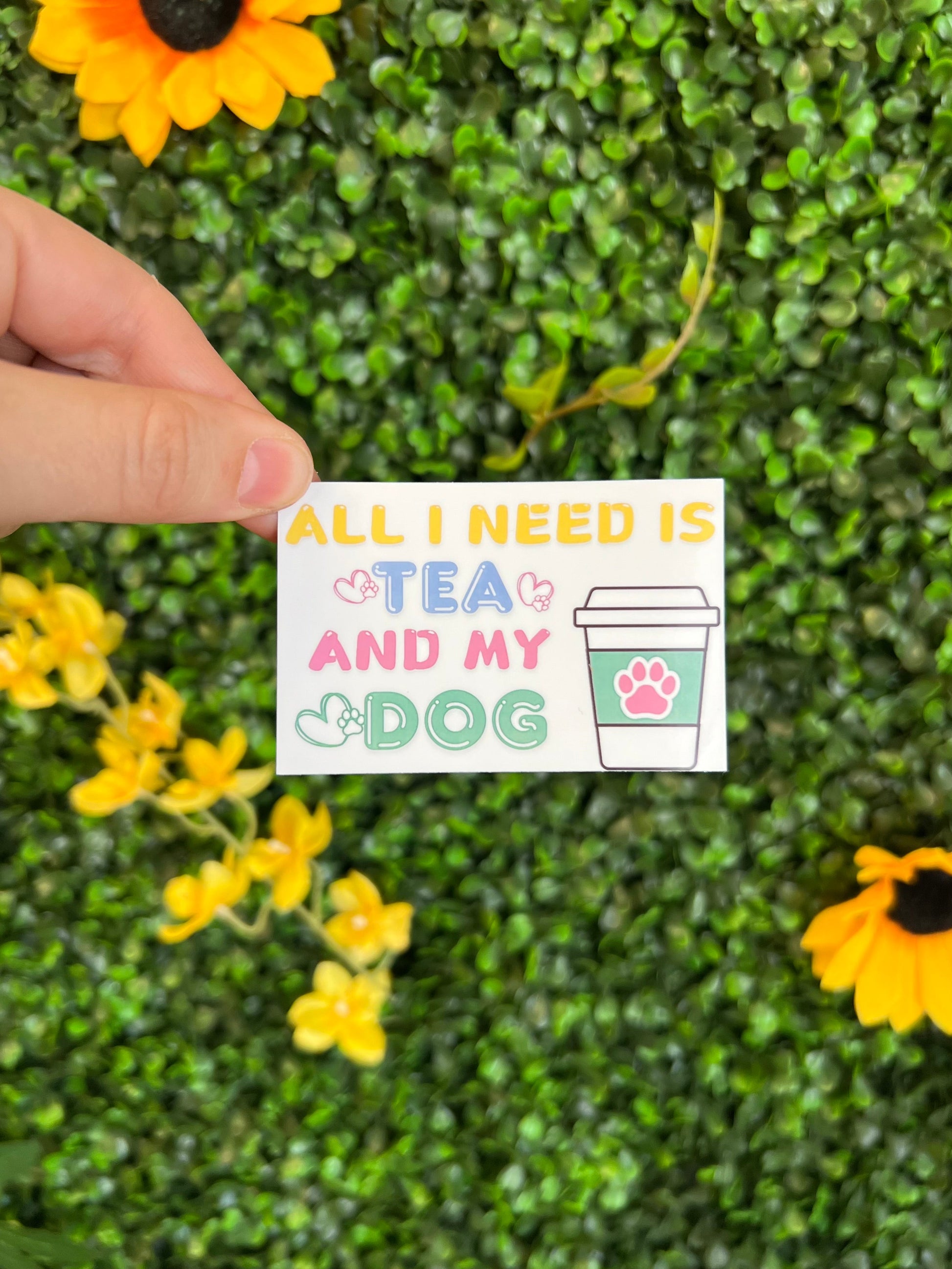 Hand holding a sticker with the phrase all I need is tea and my dog on a green leafy background